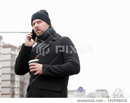 Handsome man wearing black coat and brown leather bag walking down on the city, drinking coffee paper takeaway cup with mobile phone. Stylish bearded middle-aged male using smartphone at urban street Handsome man wearing black coat and brown leather bag walking down on the city, drinking coffee paper takeaway cup with mobile phone. Stylish bearded middle-aged male using smartphone at urban street 142046038