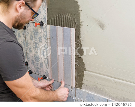 Worker Laying Ceramic Tiles on the Wall. Repairman Placing White Gray Square Tiles, Applying Adhesive. Builder Installing Tile Using Leveler, Tile Leveling Clips. Tiling. Finishing Repair Work. 4K 142046755