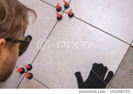 Worker Hand Laying Ceramic Tiles on the Floor. Repairman Placing White Gray Square Tiles, Applying Adhesive. Builder Installing Tile Using Leveler, Tile Leveling Clips. Tiling. Finishing Repair Work. 142046783