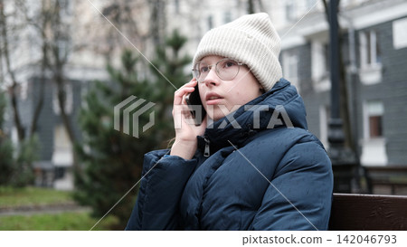 Teenager Girl Walking Alone on City Street with Smartphone. Portrait of 14 years old girl wearing eyeglasses, wool hat, winter jacket using mobile phone. Urban Style. Rainy mood weather. Cold season Teenager Girl Walking Alone on City Street with Smartphone. Portrait of 14 years old girl wearing eyeglasses, wool hat, winter jacket using mobile phone. Urban Style. Rainy mood weather. Cold season 142046793