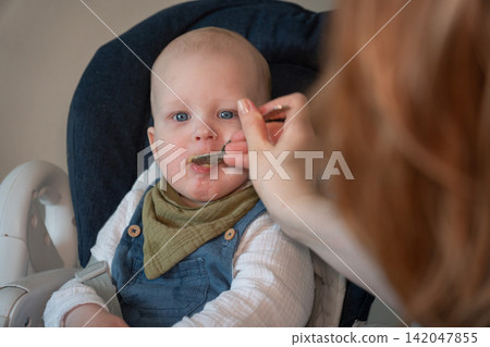 Mother brings spoonful of food toward baby sitting in high chair during mealtime at home. Infant feeding, nutrition, childcare, complementary food, parenting, healthy eating, development, family care. Mother brings spoonful of food toward baby sitting in high chair during mealtime at home. Infant feeding, nutrition, childcare, complementary food, parenting, healthy eating, development, family care. 142047855
