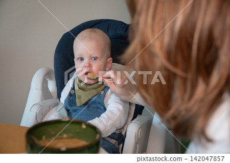 Mother feeds baby with spoon while he sits in high chair beside food bowl at home. Baby feeding, childcare, nutrition, complementary food, parenting, maternal care, development, family routine. Mother feeds baby with spoon while he sits in high chair beside food bowl at home. Baby feeding, childcare, nutrition, complementary food, parenting, maternal care, development, family routine. 142047857