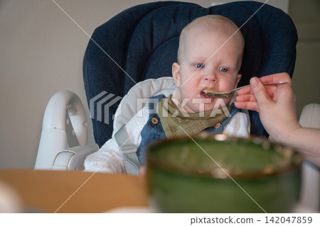 Adult feeds baby with spoon as he sits in high chair during meal at home. Baby feeding, childcare, nutrition, complementary food, healthy eating, caregiving, development, family routine. Adult feeds baby with spoon as he sits in high chair during meal at home. Baby feeding, childcare, nutrition, complementary food, healthy eating, caregiving, development, family routine. 142047859