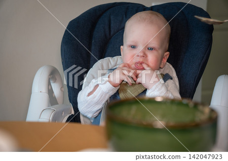 Baby watches spoon with food while sitting in high chair during mealtime at home. Baby feeding, complementary food, nutrition, childcare, curiosity, development, parenting, healthy eating. Baby watches spoon with food while sitting in high chair during mealtime at home. Baby feeding, complementary food, nutrition, childcare, curiosity, development, parenting, healthy eating. 142047923