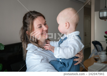 Smiling mother holds baby in her arms as they look at each other at home. Motherhood, bonding, affection, childcare, maternal love, family connection, happiness, infant development. Smiling mother holds baby in her arms as they look at each other at home. Motherhood, bonding, affection, childcare, maternal love, family connection, happiness, infant development. 142047941