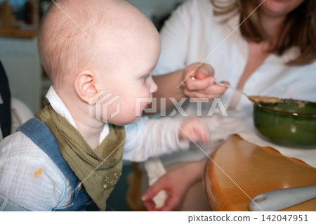 Mother feeds baby with spoon while he sits in high chair during meal at home. Baby feeding, childcare, nutrition, complementary food, parenting, maternal care, development, family routine. 142047951