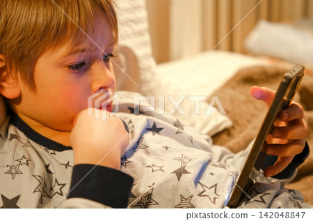 Cute kid boy in pajamas lying in bed watching smartphone, smiling. Children's screen time. Child plays games using phone at the evening or night. Social media addiction. Childhood, modern technology Cute kid boy in pajamas lying in bed watching smartphone, smiling. Children's screen time. Child plays games using phone at the evening or night. Social media addiction. Childhood, modern technology 142048847
