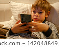 Cute kid boy in pajamas lying in bed watching smartphone, smiling. Children's screen time. Child plays games using phone at the evening or night. Social media addiction. Childhood, modern technology 142048849