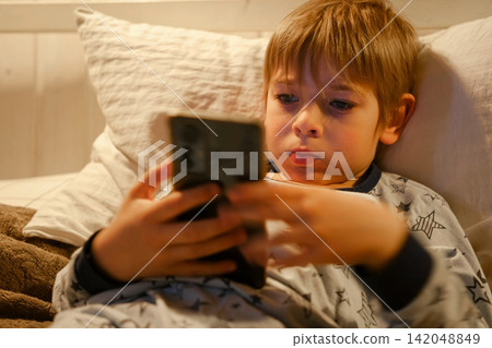 Cute kid boy in pajamas lying in bed watching smartphone, smiling. Children's screen time. Child plays games using phone at the evening or night. Social media addiction. Childhood, modern technology Cute kid boy in pajamas lying in bed watching smartphone, smiling. Children's screen time. Child plays games using phone at the evening or night. Social media addiction. Childhood, modern technology 142048849