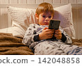 Cute kid boy in pajamas lying in bed watching smartphone, smiling. Children's screen time. Child plays games using phone at the evening or night. Social media addiction. Childhood, modern technology 142048858