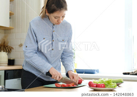 Close up of a woman chopping vegetables in a kitchen. Female hands cutting tomato, carrot on the board to preparing food. Then woman stir fried vegetables in a frying pan. Cooking in the kitchen 142048863