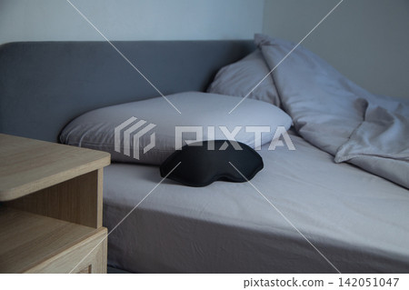 There is a black sleep mask on the bed with gray pastel linens and pillows, and part of a wooden bedside table is visible next to it. The concept: objects for sleeping in a natural home environment There is a black sleep mask on the bed with gray pastel linens and pillows, and part of a wooden bedside table is visible next to it. The concept: objects for sleeping in a natural home environment 142051047