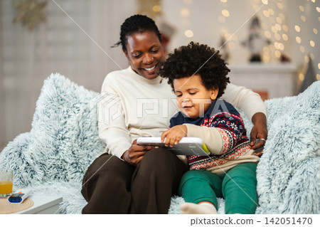 Mother and young child sitting on a sofa, happily using a digital tablet Mother and young child sitting on a sofa, happily using a digital tablet 142051470