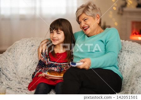 Grandmother and granddaughter laughing watching TV at home Grandmother and granddaughter laughing watching TV at home 142051501