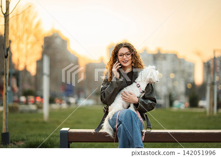 Young woman sitting on park bench talking on phone holding dog Young woman sitting on park bench talking on phone holding dog 142051996
