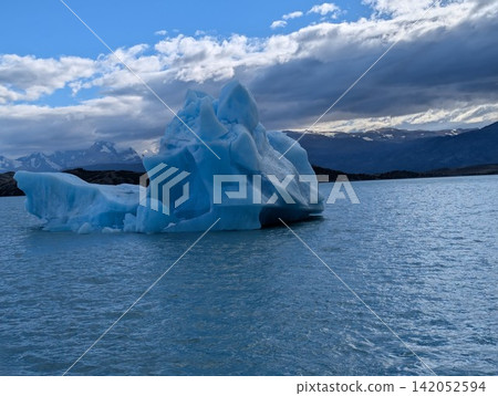 Large Blue Iceberg with Layered Ice Peaks Under Cloudy Sky 142052594
