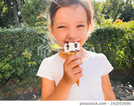 Cheerful girl smiling while eating vanilla chocolate ice cream outdoors. Happy child enjoys a sweet frozen dessert in sunlight. Childhood joy concept. Cheerful girl smiling while eating vanilla chocolate ice cream outdoors. Happy child enjoys a sweet frozen dessert in sunlight. Childhood joy concept. 142053690