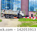 A construction dump truck is parked in front of a modern building with reflective glass windows. The truck is white with a rusted metal body and has six large tires 142053913