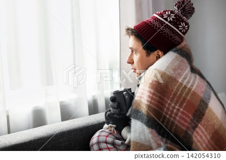 Freezing man in a winter blanket at home looks out the window with a hot mug. Cold weather and weak heating reflect the energy crisis and high costs. Freezing man in a winter blanket at home looks out the window with a hot mug. Cold weather and weak heating reflect the energy crisis and high costs. 142054310