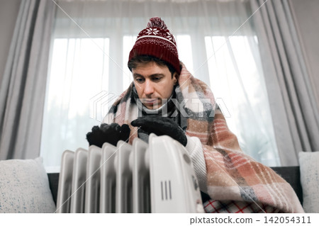 Freezing man in a winter blanket warms hands over a heater at home. Cold weather and energy crisis cause high heating costs. Freezing man in a winter blanket warms hands over a heater at home. Cold weather and energy crisis cause high heating costs. 142054311