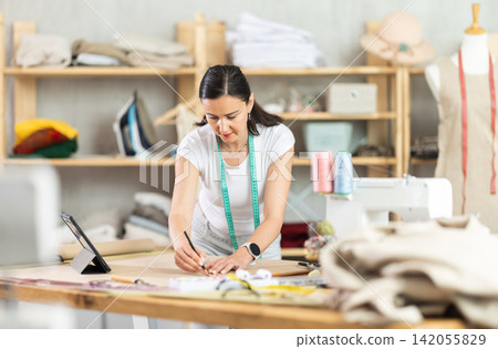 Woman tailor draws dress pattern of product on paper, tablet on table. Woman tailor draws dress pattern of product on paper, tablet on table. 142055829