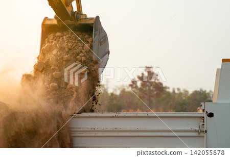 Backhoe loading soil into heavy duty dump truck at construction site. Crawler excavator transporting dirt with powerful bucket. Heavy construction machinery. Industrial earthmoving equipment in action Backhoe loading soil into heavy duty dump truck at construction site. Crawler excavator transporting dirt with powerful bucket. Heavy construction machinery. Industrial earthmoving equipment in action 142055878