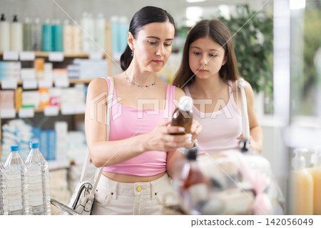 Positive family of adult woman and young girl choosing medicinal cough liquid syrup standing between shelves at pharmacy Positive family of adult woman and young girl choosing medicinal cough liquid syrup standing between shelves at pharmacy 142056049