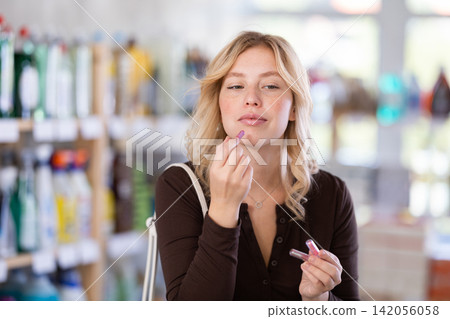 Beautiful stylish young woman choosing lipstick and trying samples in cosmetics department of hypermarket Beautiful stylish young woman choosing lipstick and trying samples in cosmetics department of hypermarket 142056058