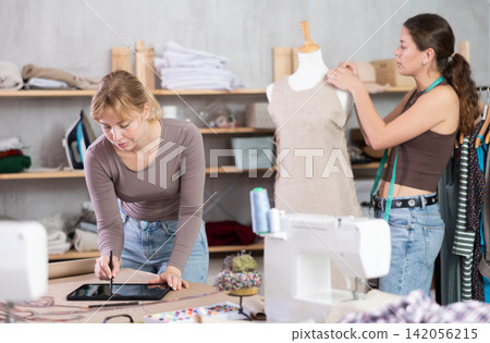 Dressmaker sketching dress on tablet while colleague working with mannequin Dressmaker sketching dress on tablet while colleague working with mannequin 142056215