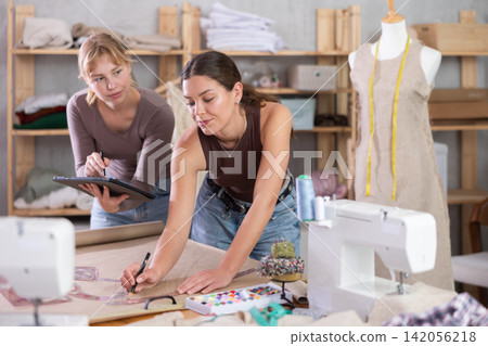 Two young female dressmakers drawing in tablet in sewing workshop Two young female dressmakers drawing in tablet in sewing workshop 142056218