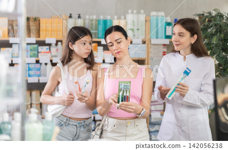 Family of adult woman and young girl choosing best toothbrush at pharmacy, young female pharmacist consulting and offering variety of goods to customers Family of adult woman and young girl choosing best toothbrush at pharmacy, young female pharmacist consulting and offering variety of goods to customers 142056238