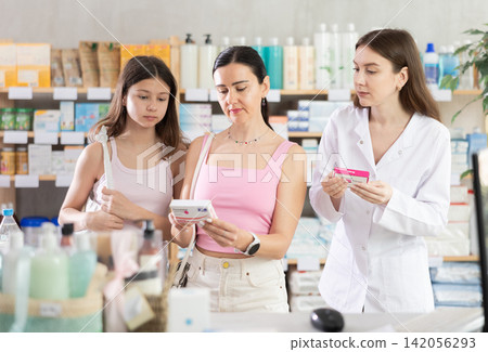 Interested mother and daughter clients looking box of paracetamol antipyretic pills, young woman pharmacist helping choosing box and making decisions in drug store Interested mother and daughter clients looking box of paracetamol antipyretic pills, young woman pharmacist helping choosing box and making decisions in drug store 142056293