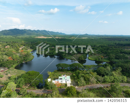 Drone view of eco friendly lakeside resort with a solar powered house, lush gardens, tropical forest and mountain scenery. Sustainable tourism, renewable energy and natural living. Responsible travel. Drone view of eco friendly lakeside resort with a solar powered house, lush gardens, tropical forest and mountain scenery. Sustainable tourism, renewable energy and natural living. Responsible travel. 142056315