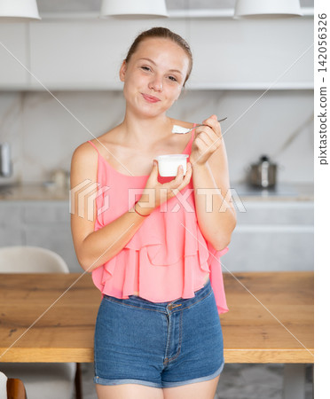 Teenage girl eating fresh yogurt with spoon in modern kitchen 142056326