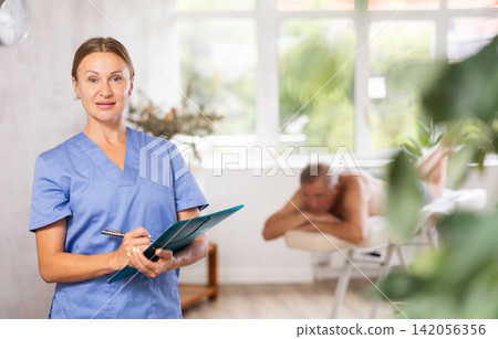 Positive middle-aged masseuse posing with folder in hand in the therapy room Positive middle-aged masseuse posing with folder in hand in the therapy room 142056356