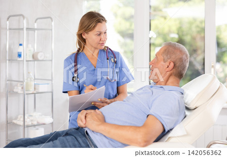 Middle-aged female doctor signing papers while talking with man patient Middle-aged female doctor signing papers while talking with man patient 142056362