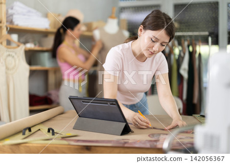 Girl tailor draws dress pattern of product on paper, tablet on table. Companion in background Girl tailor draws dress pattern of product on paper, tablet on table. Companion in background 142056367
