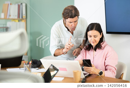 Man scolding colleague using smartphone in workplace Man scolding colleague using smartphone in workplace 142056416