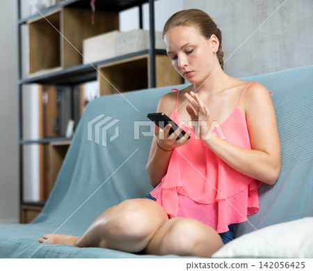 Teenager on sofa with smartphone, girl scrolls through her smartphone Teenager on sofa with smartphone, girl scrolls through her smartphone 142056425