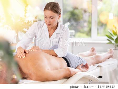 Skilled middle-aged masseuse conducting back massage for man client Skilled middle-aged masseuse conducting back massage for man client 142056458
