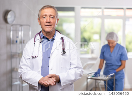 Old man doctor posing in doctor's cabinet with clinical chair Old man doctor posing in doctor's cabinet with clinical chair 142056460