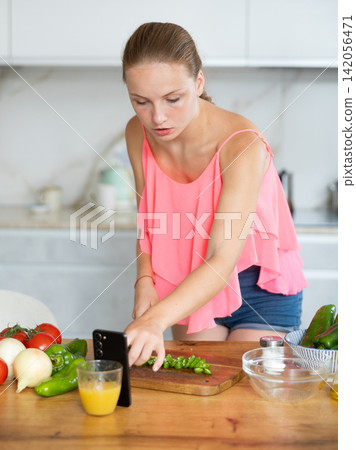 Portrait of pretty teenage girl reading recipe online on the phone in kitchen Portrait of pretty teenage girl reading recipe online on the phone in kitchen 142056471