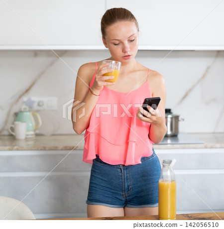Teenage girl with glass of juice scrolls through smartphone screen 142056510