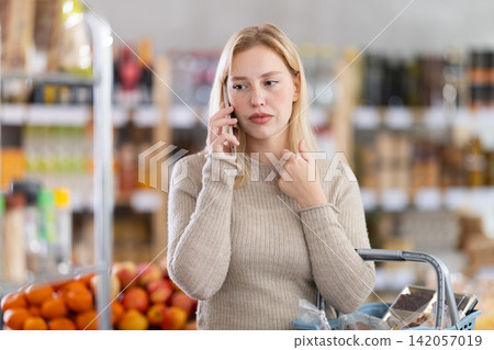 Upset young woman choosing groceries while talking on phone 142057019