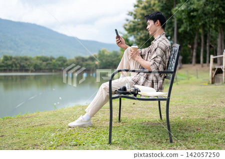 Young Asian man relaxing with phone and coffee on a bench by a scenic mountain lake in greenery park Young Asian man relaxing with phone and coffee on a bench by a scenic mountain lake in greenery park 142057250