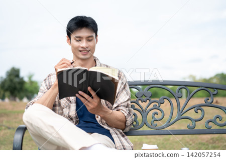Young Asian man holding pencil writing a diary book on a metal bench in park under the bright sky. 142057254