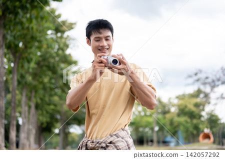 Young Asian man holding camera taking a photo while strolling through a green park under clear sky. 142057292