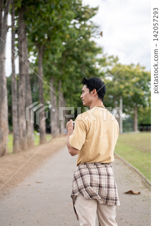 Back view shot of Young Asian man holding camera strolling along a pathway in the greenery park. Back view shot of Young Asian man holding camera strolling along a pathway in the greenery park. 142057293