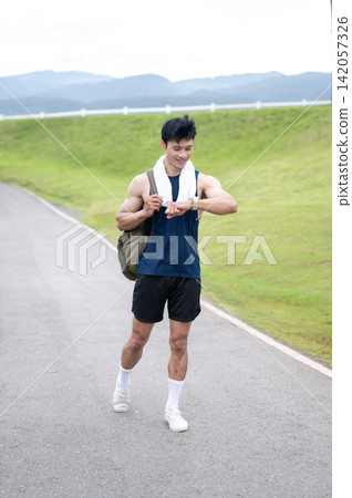 Athletic Asian man checking time on watch while walking on a quiet road surrounded by green hills. Athletic Asian man checking time on watch while walking on a quiet road surrounded by green hills. 142057326