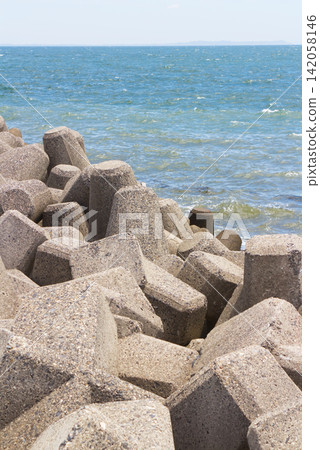 A vertical composition image featuring tetrapods piled on the coast, the blue sea, a breakwater, and an industrial landscape. A vertical composition image featuring tetrapods piled on the coast, the blue sea, a breakwater, and an industrial landscape. 142058146